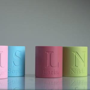 May include: Five cylindrical containers in pastel pink, light blue, dark pink, lime green, and white. Each container features a large embossed letter and a smaller engraved name, specifically Lea, Sophie, Larisa, Nikki, and Carina, on the front.