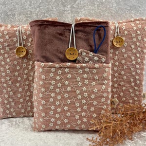 Könnte beinhalten: Drei rosa Stoffbeutel mit weißer Blumenstickerei. Die Beutel haben eine gesteppte Textur und sind mit Holzknöpfen und weißen Schnurschlaufen verziert. Einer der Beutel hat ein braunes Samtinnenfutter und ein kleines passendes Accessoire.