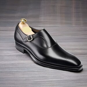May include: A black leather single monk strap shoe with a silver-toned metal buckle, a pointed toe featuring subtle decorative perforations, and a wooden shoe tree inserted inside the heel area to maintain the shape of the footwear.