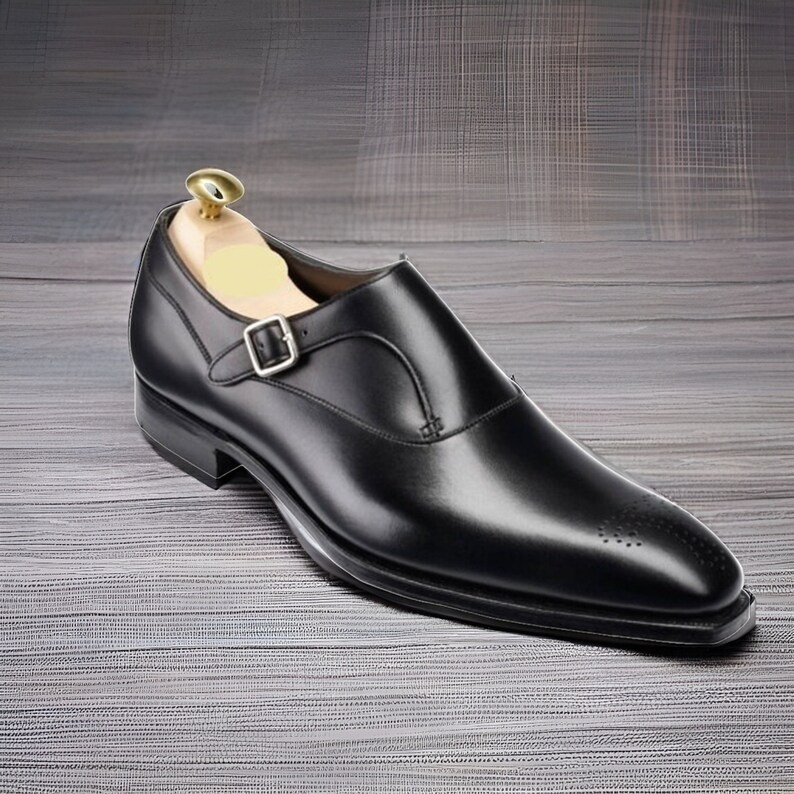 May include: A black leather single monk strap shoe with a silver-toned metal buckle, a pointed toe featuring subtle decorative perforations, and a wooden shoe tree inserted inside the heel area to maintain the shape of the footwear.