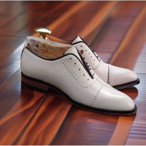 May include: A pair of white leather dress shoes with black trim and decorative perforations. The shoes have a classic Oxford design with a low heel and are displayed on a wooden surface.