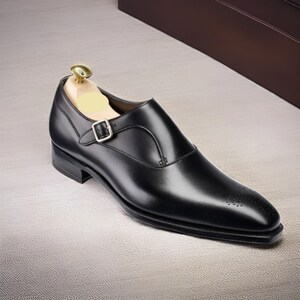 May include: A black leather single monk strap shoe with a silver-toned buckle and a wooden shoe tree inserted. The shoe features a pointed toe with subtle decorative perforations along the front edge and a low stacked heel.