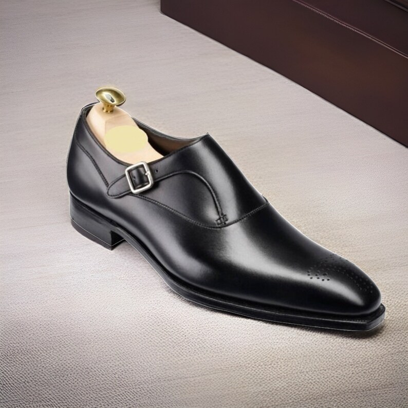 May include: A black leather single monk strap shoe with a silver-toned buckle and a wooden shoe tree inserted. The shoe features a pointed toe with subtle decorative perforations along the front edge and a low stacked heel.