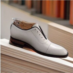May include: White leather shoe with a black trim and a brown heel. The shoe has a pointed toe and decorative perforations along the side. The shoe is a size 9.