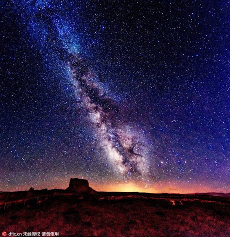 May include: A night sky with a vast expanse of stars, including the Milky Way galaxy, stretching across the horizon. The sky is a deep blue with a hint of purple, and the stars are bright and numerous. A dark silhouette of a rock formation is visible in the foreground.