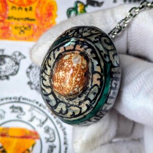 Puede incluir: Colgante ovalado con una concha central moteada marrón y blanca, rodeada por un patrón decorativo en negro, gris y verde. El colgante está sujeto a una cadena plateada, mostrando un diseño único y artesanal.
