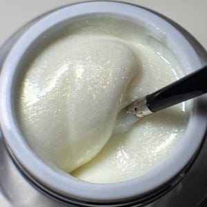 Puede incluir: Un esmalte de uñas de gel blanco y brillante con un pincel sumergido en el producto. El gel está en un recipiente transparente con una tapa blanca.
