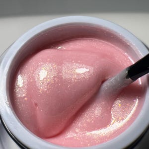 Puede incluir: Primer plano de un esmalte de uñas de gel rosa con un acabado brillante. El esmalte está en un frasco transparente con un borde blanco. Un pincel está parcialmente sumergido en el esmalte.