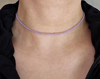 Könnte beinhalten: Eine zarte, lavendelfarbene Perlen-Choker-Halskette. Die Halskette hat kleine, runde Perlen in einem sanften Lila-Ton, mit einem Abschnitt aus silberfarbenen Perlen. Die Halskette wird um den Hals getragen.