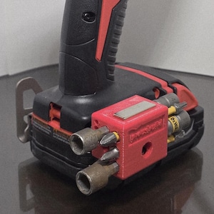 May include: A black and red cordless drill with a red bit holder attached. The bit holder has several bits and a magnetic plate. The drill is on a reflective surface.