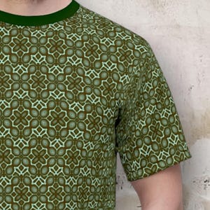 May include: Green short-sleeved t-shirt with a repeating geometric pattern in shades of green and brown. The shirt has a dark green crew neck. The pattern is a series of interconnected floral-like shapes.