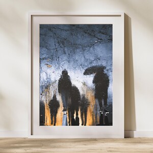 May include: Silhouettes of people walking in the rain, reflected in a puddle on a city street. The image is in black and white, with a blue-grey background.