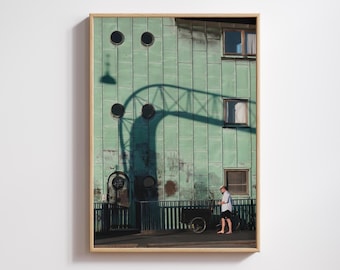 May include: Framed photograph of a building with a weathered, teal-green facade. The building has circular windows and rectangular windows. A person walks along the sidewalk next to a small cart. The shadow of a structure arches across the building.