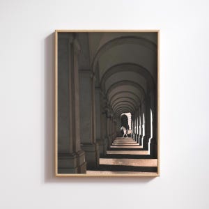 May include: Framed art print featuring a long, arched corridor with repeating columns and a figure in the distance. The artwork has a warm color palette with shadows and sunlight creating a dramatic effect. The frame is a light wood color.