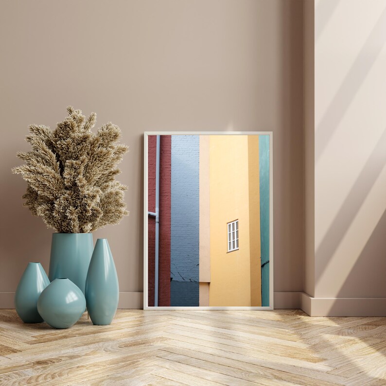 May include: A framed print of an abstract cityscape with vertical stripes of red, blue, yellow, and green. The print features a single window in the yellow stripe.
