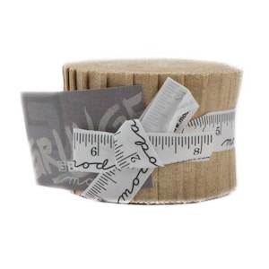 May include: A tan fabric roll is encircled by a white ribbon printed with ruler markings and text. A grey tag with the text "Girl Brod" is attached. The fabric roll is approximately 15 cm in diameter.