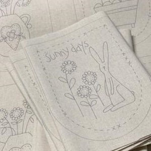 May include: Pile of light beige fabric napkins with a hand-drawn design. Each napkin features a bunny, flowers, and the words "sunny days" in an oval. Other napkins and floral designs are visible in the background.