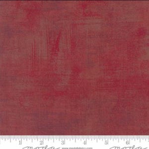 May include: Red fabric with a textured, mottled appearance. The fabric has a distressed look with vertical and horizontal brushstroke-like patterns. A ruler is at the bottom of the fabric. The fabric is likely used for quilting or other craft projects.