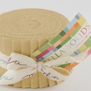 May include: A roll of tan fabric strips, secured with a cream ribbon bow. The ribbon features blue and brown script. A colourful tag reads "BELLA SOLIDS moda". The fabric is tightly wound, showing the edges of the strips.