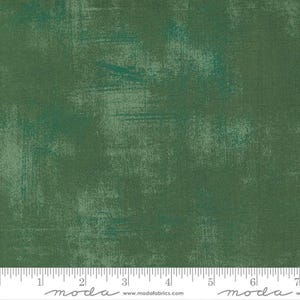 May include: Green fabric with a textured, distressed appearance. The fabric has a mottled effect with lighter green brushstrokes over a darker green base. A ruler is at the bottom of the image, with measurements in inches.