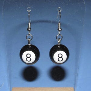 May include: A pair of black and white 8-ball earrings. Each earring features a black sphere with a white circle and the number 8. The earrings hang from silver-colored fish hook ear wires.
