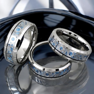 May include: Three silver tungsten carbide rings with a beveled edge and a blue and silver patterned design.