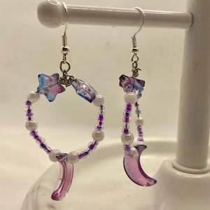 May include: A pair of handmade earrings with a celestial theme. One earring is a hoop with white and purple beads, a pink crescent, and star-shaped accents. The other earring is a dangling design with similar elements.