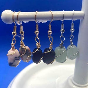 Peut inclure: Présentation de boucles d'oreilles en pierres précieuses faites à la main. Les boucles d'oreilles présentent diverses pierres, notamment roses, noires et vertes, chacune enveloppée de fil d'or ou d'argent. Elles sont suspendues à un présentoir blanc sur fond bleu.