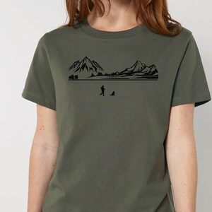 Può includere: T-shirt verde oliva con grafica nera raffigurante una catena montuosa, una persona e un cane. Il disegno è centrato sul petto. La maglietta ha maniche corte e girocollo.