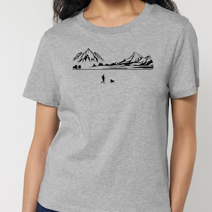 Puede incluir: Camiseta gris con un gráfico negro de una persona paseando a un perro frente a una cordillera. El diseño incluye montañas, árboles y un lago. La camiseta tiene mangas cortas y cuello redondo.