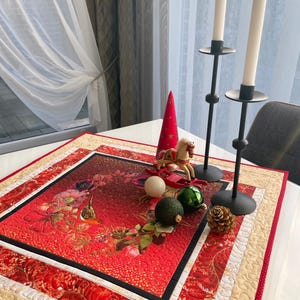 Könnte beinhalten: Eine festliche Weihnachtsdekoration mit einem rot-goldenen, gesteppten Tischläufer mit floralen Mustern. Enthält ein Holzschaukelpferd, Ornamente, eine Tannenzapfen und zwei schwarze Kerzenhalter mit weißen Kerzen. Eine rote Kegeldekoration ist ebenfalls vorhanden.