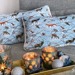 Può includere: Due cuscini decorativi con una stampa di scena invernale azzurra con cervi, volpi, gufi e scoiattoli. I cuscini sono abbinati a portacandele illuminati e ornamenti decorativi su un vassoio dorato.