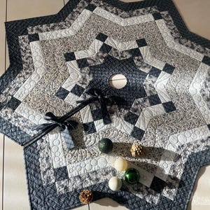 Könnte beinhalten: Ein sternförmiger, gesteppter Baumrock in Grau-, Weiß- und Schwarztönen. Das Design weist geometrische Muster und florale Akzente auf. Dekorative Ornamente und eine Schleife sind auf dem Rock platziert.