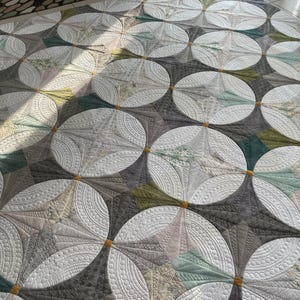 May include: A large, handmade quilt with a repeating pattern of overlapping circles. The circles are mainly white with detailed quilted designs, set against a backdrop of grey, green, and tan fabrics. This decorative home textile is a quilt.