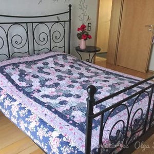 Könnte beinhalten: Ein schwarzes Schmiedeeisenbett mit einer floralen Steppdecke. Die Steppdecke hat ein Patchwork-Design in Blau-, Rosa- und Grautönen. Das Bett steht in einem Schlafzimmer mit Holzboden und einer weißen Kommode.