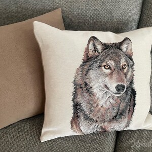 Op de afbeelding: Een decoratief sierkussen met een gedetailleerd portret van een wolf. De wolf heeft grijze en bruine vacht, met gele ogen. Het kussen staat op een neutrale achtergrond, met een tweede effen gekleurd kussen op de achtergrond.