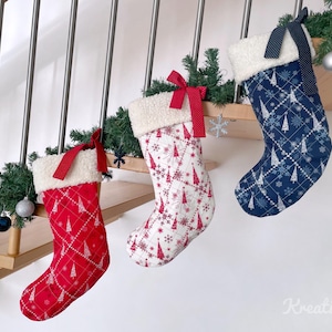 May include: Three Christmas stockings in red, white, and blue, each with a quilted pattern of trees and snowflakes. They have a fluffy white cuff and a ribbon for hanging. The stockings are displayed on a wooden staircase.