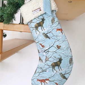 May include: A Christmas stocking with a light blue background featuring a winter forest scene with deer, owls, foxes, and squirrels. The top is white and fluffy, with a light blue bow. The stocking is hanging on a wooden staircase.