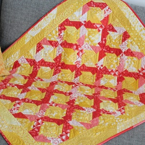 Könnte beinhalten: Eine handgefertigte Steppdecke mit einem geometrischen Muster in Gelb-, Rot- und Weißtönen. Die Steppdecke weist ein sich wiederholendes Design aus ineinandergreifenden Formen mit einem gelben Rand und einer rosa Bindung auf. Ein dekoratives Kissen mit einem orangefarbenen Blumenmuster ist ebenfalls sichtbar.