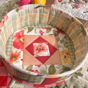 May include: A round, quilted fabric basket with a floral and plaid pattern. The interior features a central square with a red and peach floral design. The rim is lined with a green and yellow plaid fabric. The basket is on a floral patterned surface.