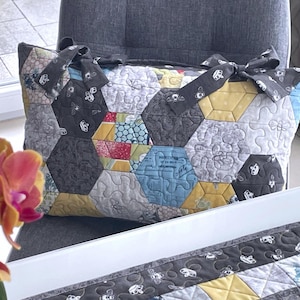 Puede incluir: Un bolso tote acolchado con un patrón geométrico de hexágonos en varios colores, incluyendo gris, amarillo, azul y rojo. El bolso tiene lazos grises en las asas. Se ve un mantel individual acolchado a juego.
