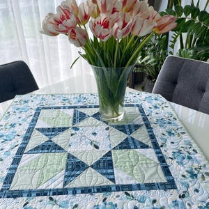 Op de afbeelding: Een vierkante gewatteerde tafelloper met een bloemen- en geometrisch patroon in blauw- en groentinten. Een vaas met tulpen met rood-wit gestreepte bloemblaadjes staat op de loper. De loper ligt op een witte tafel.