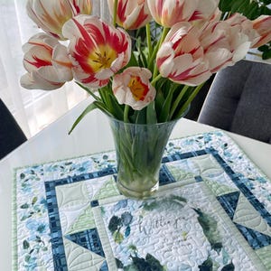 Könnte beinhalten: Ein Strauß weiß-rot gestreifter Tulpen in einer durchsichtigen Glasvase steht auf einem weißen Tisch. Darunter befindet sich ein gesteppter Tischläufer mit blauen Blumen- und Karomustern. Auf dem Tischläufer steht der Text "Within you..."