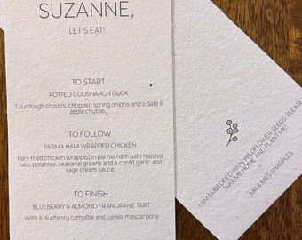 May include: A white card with a handwritten menu for a dinner party. The menu includes a starter, main course, and dessert. The card is decorated with a floral illustration.