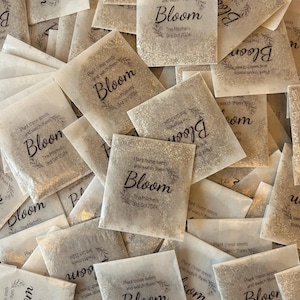 Op de afbeelding: Een stapel kleine, witte papieren zakjes gevuld met zaden. Elk zakje heeft een bruin label met de tekst "Plant deze zaden en bekijk hoe ze bloeien. De Mitchells. 3 oktober 2024."