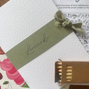 May include: A white card with a green ribbon and the name "Leonardo" written on it. The card is on a wooden table with a set of coloured pencils and a sheet of paper with a wedding-themed colouring page.