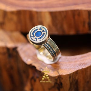 Peut inclure: Une bague dorée avec un motif circulaire en émail bleu. La bague présente des détails complexes sur le bandeau et un emblème bleu central. La bague est posée sur un fond en bois, soulignant son savoir-faire.
