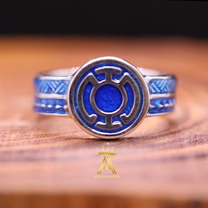Pode incluir: Um anel de prata com um emblema circular azul no centro. O emblema tem uma contorno prateado e um centro azul. A banda do anel tem detalhes azuis. Colocado sobre uma superfície de madeira.
