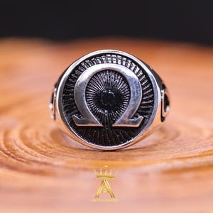 Könnte beinhalten: Ein silberner Ring mit dem Omega-Symbol. Die runde Oberfläche des Rings hat ein strukturiertes Muster, das vom Zentrum ausgeht. Das Band weist dekorative Details auf. Der Ring wird auf einer Holzoberfläche präsentiert.