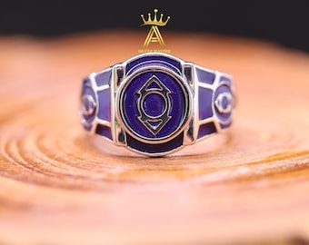 May include: A silver-toned ring featuring a central circular emblem with a purple enamel inlay and a stylized arrow-like symbol. The band is decorated with additional purple enamel panels and smaller eye-shaped motifs on the sides of the ring.